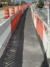 Channelizing devices guide pedestrians away from the existing sidewalk while crews place new sidewalk along the east side of the roadway. February 2026